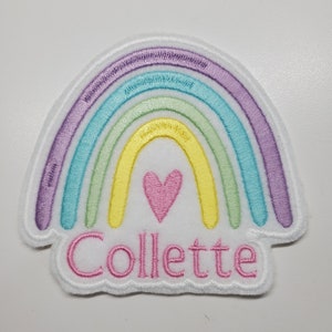 May include: A white felt rainbow patch with a pink heart in the center and the name "Collette" embroidered in pink below.