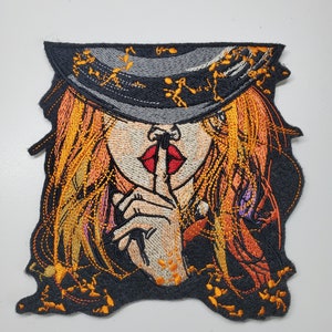 Peut inclure: Patch brodée thermocollante représentant une femme aux longs cheveux blonds portant un chapeau noir et un rouge à lèvres rouge. Elle se tient le doigt sur les lèvres, comme pour dire "chut". Le fond est noir avec des accents orange et or.