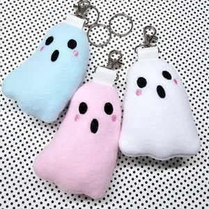 May include: Three plush ghost keychains in pastel colors: light blue, pink, and white. Each ghost has black eyes and mouth, with pink blush. They have silver key rings and white leatherette straps.