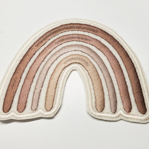 Puede incluir: Un parche bordado de arcoíris de color marrón y beige sobre un fondo blanco.