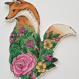 Könnte beinhalten: Gestickter Aufnäher mit einem Fuchs und einem Blumendesign. Der Fuchs ist orange und weiß, die Blumen sind rosa, gelb und grün.