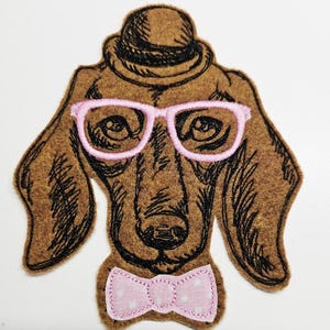 May include: A brown felt dog applique with a black outline, wearing a brown hat, pink glasses, and a pink polka-dot bow tie. The dog's face is detailed with embroidered features. This applique is suitable for crafts and sewing projects.
