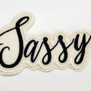 Op de afbeelding: Witte vilt patch met zwarte geborduurde tekst die "Sassy" luidt.