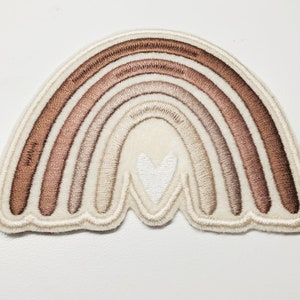 Könnte beinhalten: Ein beigefarbener Filz-Regenbogen-Patch mit einem weißen Herz in der Mitte. Der Regenbogen hat fünf Bänder in Braun, Beige und Weiß.