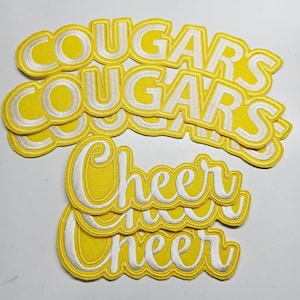 Pode incluir: Patches bordados amarelos com letras brancas. Os patches apresentam as palavras "COUGARS" e "Cheer". Estes patches são provavelmente para roupas desportivas ou artigos de apoio.