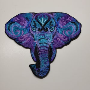 Può includere: Toppa ricamata a forma di elefante nei toni del blu e del viola. La testa dell'elefante presenta intricati motivi a spirale e dettagli decorativi. La toppa ha un bordo nero ed è progettata per essere applicata su tessuto.