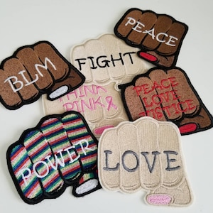 May include: A collection of embroidered fist patches in various colors and designs. The patches feature words like "BLM", "FIGHT", "PEACE", "LOVE", and "POWER". Some fists are brown, beige, or striped with rainbow colors. The patches are on a white surface.