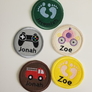 May include: Five embroidered felt circles with different designs and names. The designs include a video game controller, a carriage, a fire truck, and two sets of footprints. The names are Jonah, Zoe, and Jeremiah.