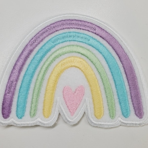 Puede incluir: Un parche bordado con forma de arcoíris con un corazón rosa en el centro. El arcoíris tiene cinco colores: morado, azul, verde, amarillo y blanco.
