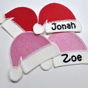 May include: Four felt Christmas hats with white trim and pom-poms. Two hats are red and two are pink. The hats are personalised with the names "Jonah" and "Zoe".