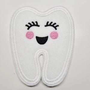 May include: A white, tooth-shaped patch with a smiling face. The tooth has black eyelashes, pink cheeks, and a black mouth. The patch is outlined with white stitching.