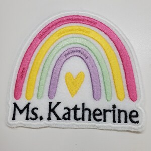 Könnte beinhalten: Gestickter Aufnäher mit einem Regenbogen-Design in Rosa, Gelb, Grün und Lila sowie einem gelben Herz. Der Aufnäher hat den Text "Ms. Katherine" in Schwarz und einen weißen Rand.