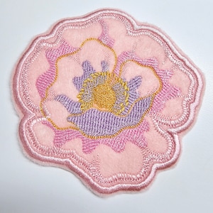 May include: A pink felt flower patch with a purple and yellow embroidered center. The flower has a scalloped edge.