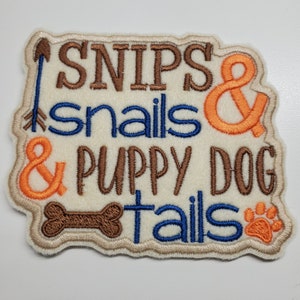 May include: A beige fabric patch with brown, orange, and blue embroidered text that reads "Snips & snails & puppy dog tails". The patch features a brown bone and an orange paw print.