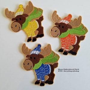 May include: Three embroidered moose patches. Each moose has brown antlers, a brown body, and a smiling face. They wear different colored sweaters, hats, and green scarves. The text "Moose Embroidered Patch" and "ETSY: thecuttingcafeshop" is visible.