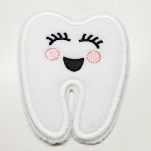 May include: A white felt tooth-shaped patch with embroidered details. The tooth has a smiling face with black eyelashes, a black mouth, and pink cheeks. The edges are outlined with white stitching.
