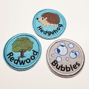 Peut inclure: Trois patchs brodés thermocollants. L'un est bleu avec un arbre vert et le texte "Redwood". L'autre est bleu avec un hérisson brun et le texte "Hedgehog". Le dernier est gris avec des bulles bleues et le texte "Bubbles".