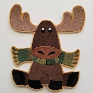 以下が含まれることがあります： 大きな角を持つ茶色のヘラジカが、緑と茶色のストライプの入ったマフラーをしています。ヘラジカは足を組んで座っており、友好的な表情をしています。