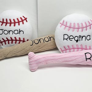 Könnte beinhalten: Zwei Plüsch-Baseballs mit den gestickten Namen "Jonah" und "Regina" und passende Plüsch-Schläger mit den gleichen Namen.