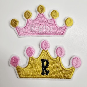 Puede incluir: Dos parches bordados con forma de corona. Uno es rosa con el nombre "Regina" bordado. El otro es dorado con la letra negra "R". Ambas coronas tienen un borde blanco y detalles dorados o rosas.