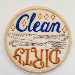 May include: A white round felt patch with the words "Clean Dish" embroidered in orange and "Clean" embroidered in blue. The patch has a decorative border.