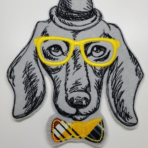 May include: Grey felt dog applique with a black sketched design, yellow glasses, and a bow tie. The dog wears a black hat. The bow tie has a plaid pattern with yellow, red, and blue accents. The glasses are a bright yellow colour.
