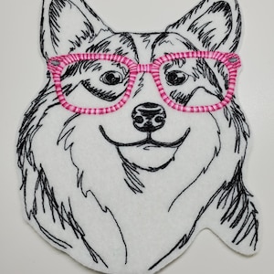 以下が含まれることがあります： ピンクの眼鏡をかけた犬の刺繍パッチ。犬は白い背景に黒い糸で描かれています。