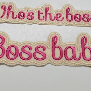 Pode incluir: Dois patches bordados de cor rosa sobre um fundo branco. O patch superior diz "Who's the boss?" e o patch inferior diz "Boss baby".