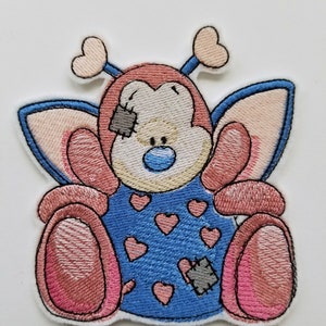 Pode incluir: Etiqueta bordada para passar a ferro com um urso de desenho animado rosa e azul com asas, que usa um fato azul com corações e uma bandagem.