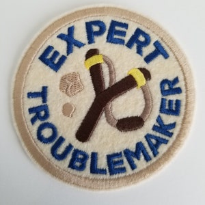 Puede incluir: Un parche redondo bordado de color beige con texto azul que dice "Expert Troublemaker". El parche presenta una honda marrón con bandas amarillas.
