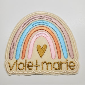 Könnte beinhalten: Ein beigefarbener Filz-Regenbogen-Patch mit einem goldenen Herzen in der Mitte und dem in Gold gestickten Text "violet + marie".