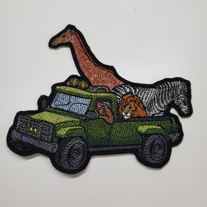 May include: An embroidered patch of a green jeep with a giraffe, zebra, and lion inside. The jeep has black wheels and yellow headlights.