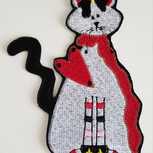 May include: Embroidered cat patch with a white body, black tail, and head with black accents. The cat wears a red scarf and has a star-shaped crown. The design includes a stylized building with red, black, and pink details, and yellow stars.