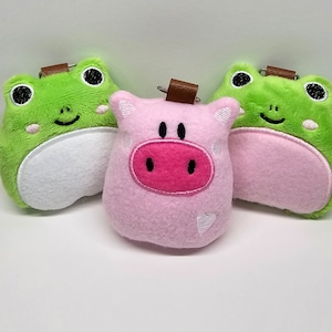 May include: Three plush toys: a green frog, a pink pig, and another green frog. Each toy has a loop for hanging.
