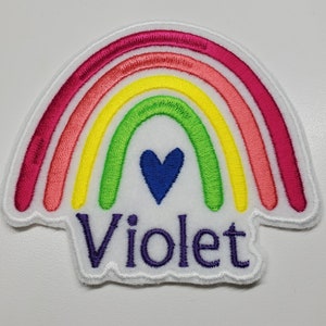 Puede incluir: Un parche de fieltro blanco en forma de arcoíris con un corazón azul en el centro y el nombre "Violet" bordado en morado debajo.