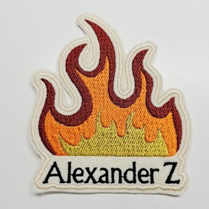 Puede incluir: Parche bordado con forma de llamas, con tonos de rojo, naranja y amarillo. El parche tiene un borde blanco y el texto "Alexander Z" en negro.
