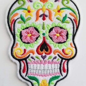 May include: A colorful embroidered sugar skull patch with pink flowers, green leaves, and yellow accents. The skull is white with black outlines and a red mouth.