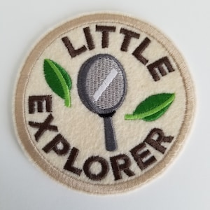 May include: A beige, round, embroidered patch with a brown border. The patch features a magnifying glass, two green leaves, and the text "LITTLE EXPLORER".