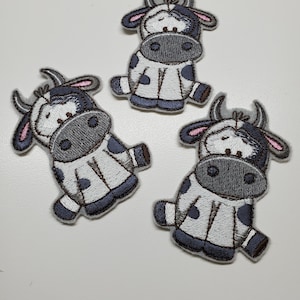 Op de afbeelding: Drie geborduurde patches in de vorm van een cartoonkoe. De patches hebben een wit lichaam met zwarte vlekken, een grijs gezicht en zwarte en roze accenten. De koe zit met uitgestrekte armen en benen.