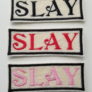 Puede incluir: Tres parches de tela blanca con bordes negros. Cada parche tiene la palabra "SLAY" bordada en hilo negro, rojo y rosa.
