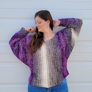 May include: A purple and brown ombre crochet sweater with a loose fit and long sleeves. The sweater is made with a soft, textured yarn.