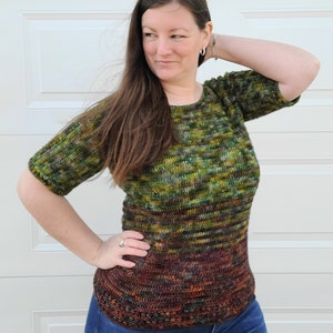 May include: A green, brown, and orange crochet top with a striped pattern. The top has a round neckline and short sleeves. The model is wearing blue jeans.