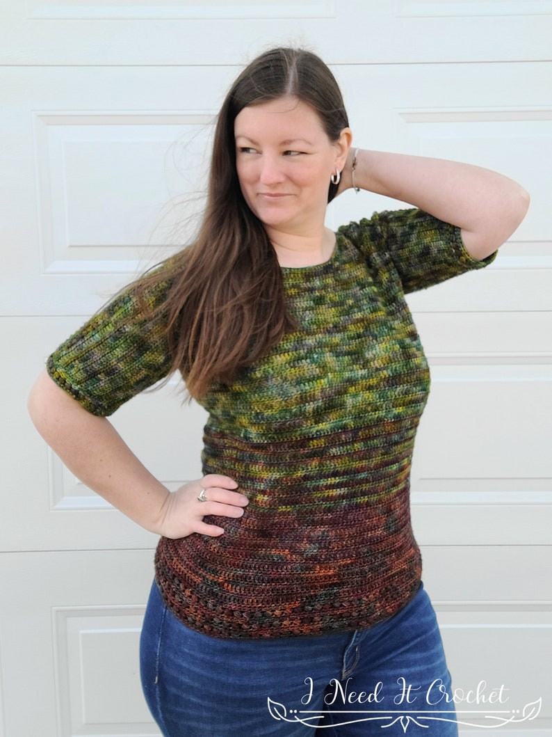 May include: A green, brown, and orange crochet top with a striped pattern. The top has a round neckline and short sleeves. The model is wearing blue jeans.