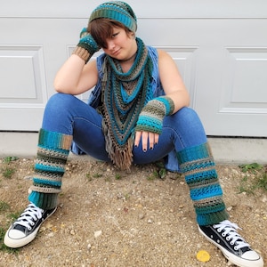 Peut inclure: Une personne portant un chapeau, une écharpe, des mitaines et des jambières en crochet bleu et vert. La personne est assise sur une surface de gravier portant un jean bleu et des baskets noires.