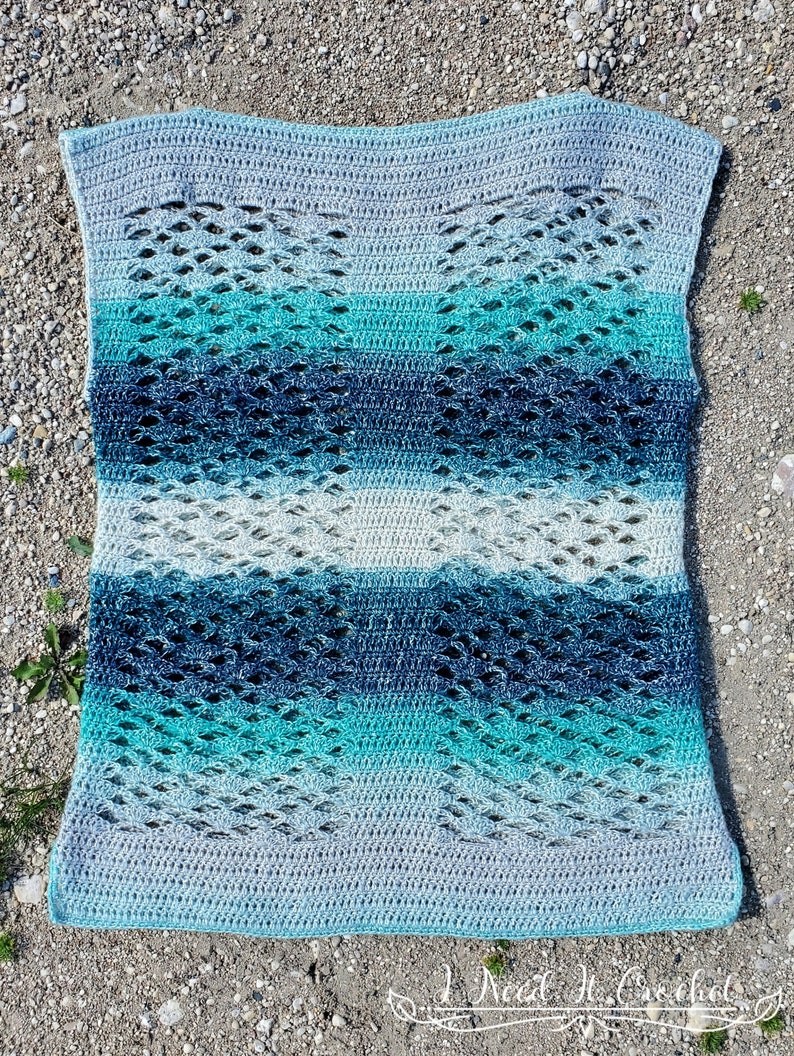 May include: A crocheted blanket with a lacy pattern in shades of blue and grey. The blanket is made with a gradient yarn that transitions from light grey to dark blue.