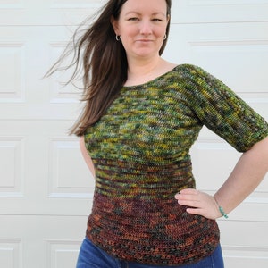May include: A green, brown, and orange crochet top with a loose fit and short sleeves. The top has a gradient pattern and is worn with blue jeans.