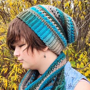 Puede incluir: Una persona lleva un gorro de punto de color azul, verde y gris a rayas y una bufanda a juego. El gorro es holgado y la bufanda tiene flecos largos. La persona está de pie frente a un fondo de hojas amarillas.