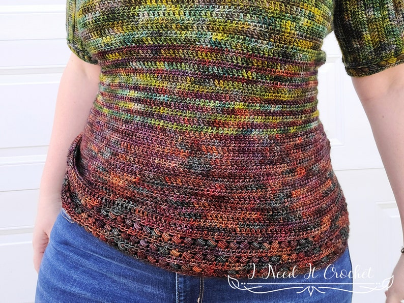 May include: A close-up of a crocheted top with a colorful, variegated yarn in shades of green, brown, and orange. The top has a fitted bodice and a wide, flared hem. The text "I Need It Crochet" is visible in the lower right corner of the image.