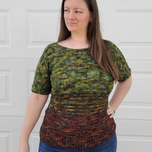 May include: A green, brown, and yellow striped crochet top with short sleeves. The top has a darker brown band at the bottom. The model is wearing blue jeans.