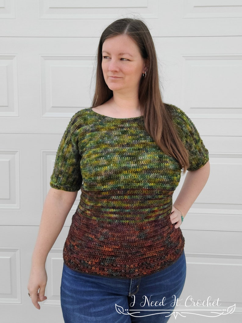 May include: A green, brown, and yellow striped crochet top with short sleeves. The top has a darker brown band at the bottom. The model is wearing blue jeans.